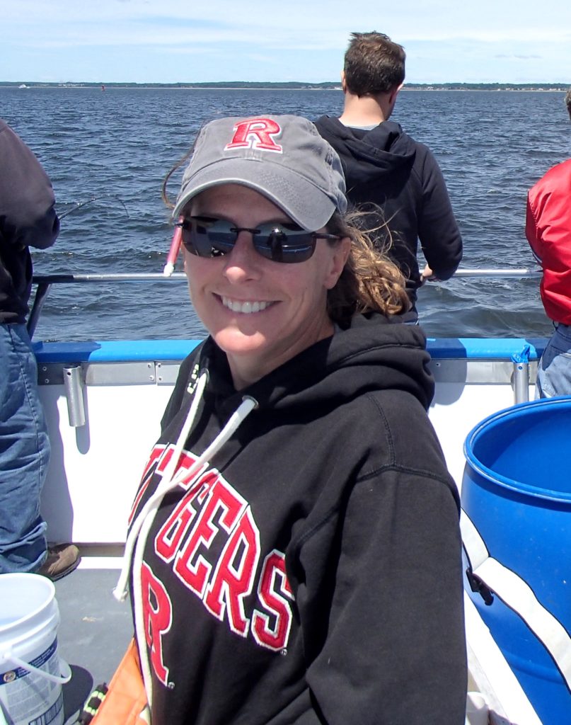 Daphne with her hair pulled back, wearing sunglasses, a gray Rutgers hat, and a black Rutgers sweatshirt. She is on a boat with others and smiling at the camera.