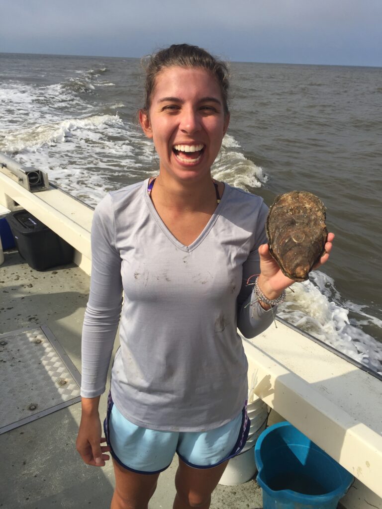 A photo of Emily, wearing a gray shirt and shorts on a boat. She is smiling widely and holding an oyster.